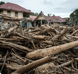 Debris and fallen trees piled up against damaged houses after a natural disaster.