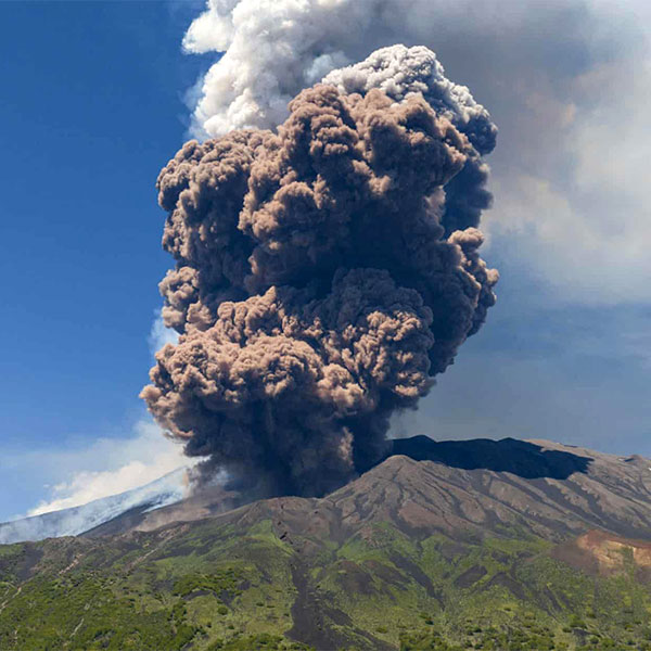 A volcano erupting, spewing thick clouds of ash and smoke into the sky above a mountainous landscape.