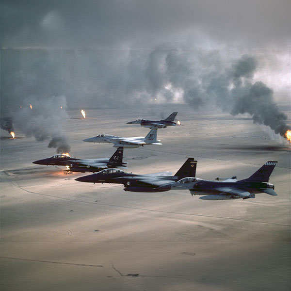 Aerial view of multiple military fighter jets flying over a desert landscape with smoke and flames in the background.
